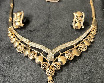 May include: A gold-toned jewelry set featuring a necklace, ring, and earrings. The necklace has a V-shaped design with a pendant. The ring and earrings have matching floral details. The set is embellished with clear crystals.