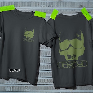 以下が含まれることがあります： チャコールグレーのTシャツ。左胸にゴーグルを着用したスカルの小さなオリーブグリーンのグラフィック、背面に同じグラフィックの大きなバージョンがプリントされており、どちらもオリーブグリーンの輪郭フォントでG59というテキストが添えられています。