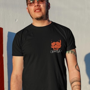 May include: A man wearing a black t-shirt with a red skull and chains graphic print. He is wearing blue jeans and has a tattoo on his arm.