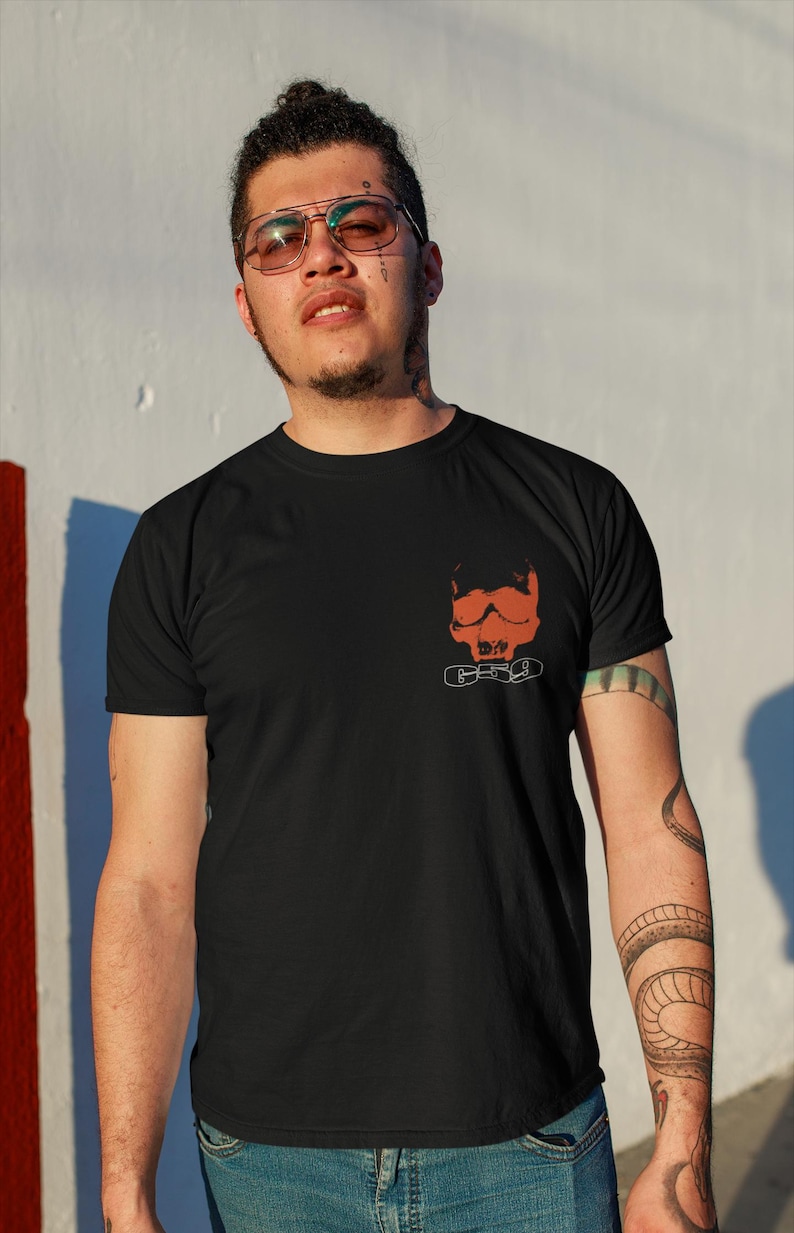 May include: A man wearing a black t-shirt with a red skull and chains graphic print. He is wearing blue jeans and has a tattoo on his arm.