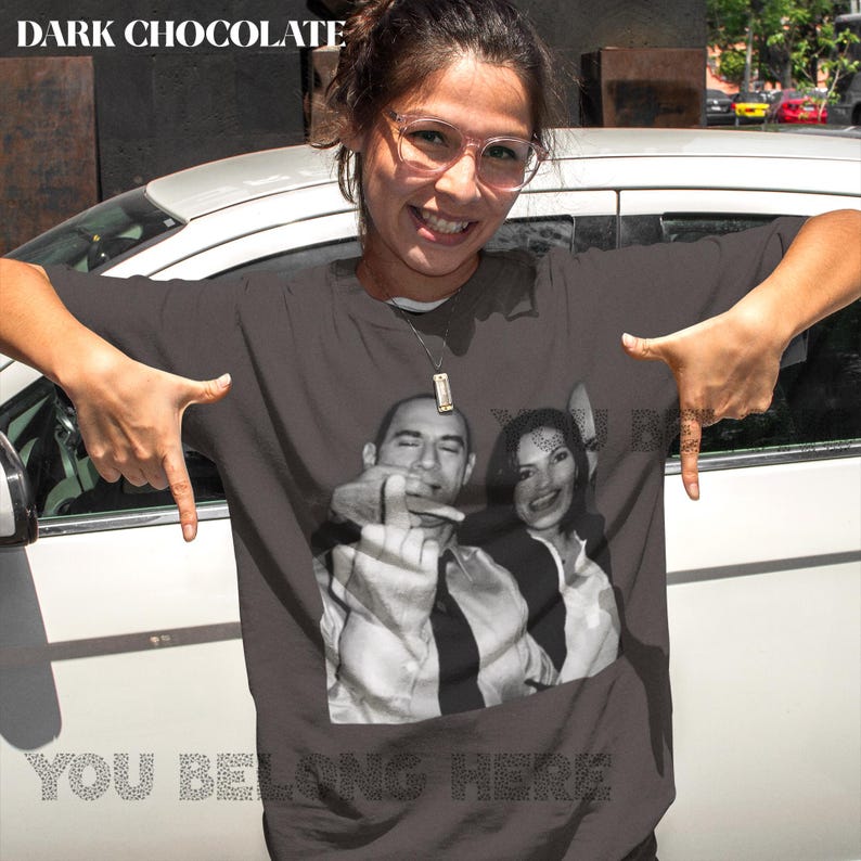 May include: A dark gray t-shirt with a black and white photo of two people. The photo shows a man and a woman with their middle fingers raised. The text "You Belong Here" is printed in white below the photo.