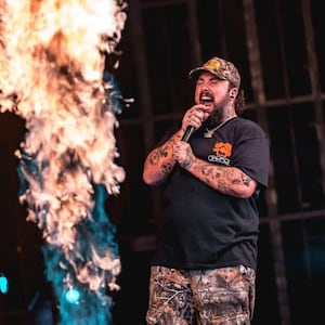 May include: A man with a beard and tattoos is singing into a microphone on stage. He is wearing a black t-shirt with a logo, camouflage shorts, and a brown and yellow baseball cap. There is a large flame behind him.