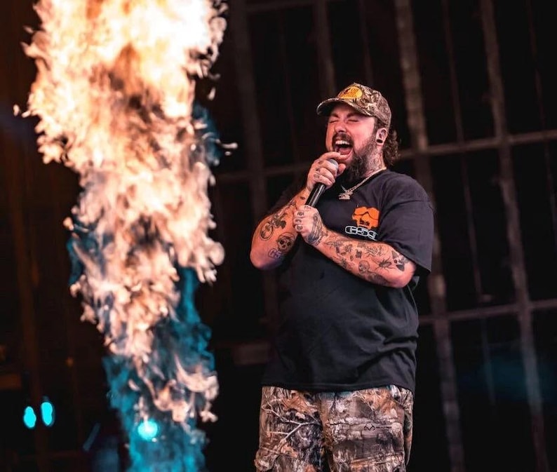 May include: A man with a beard and tattoos is singing into a microphone on stage. He is wearing a black t-shirt with a logo, camouflage shorts, and a brown and yellow baseball cap. There is a large flame behind him.