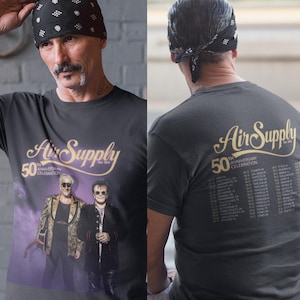 Puede incluir: Camiseta gris oscuro con el nombre de la banda Air Supply en letras doradas. La parte delantera de la camiseta muestra el texto "50th Anniversary Celebration" y una foto de la banda. La parte trasera de la camiseta enumera las fechas de la gira. Un artículo musical.