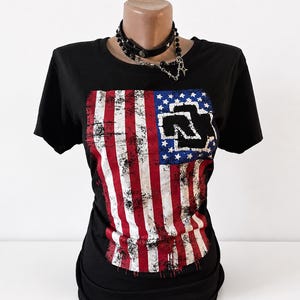 Puede incluir: Una camiseta negra de manga corta con un gr&aacute;fico de la bandera estadounidense desgastada en la parte delantera y el logotipo negro de Rammstein sobre el campo de estrellas azul. La prenda se muestra en un maniqu&iacute; con una gargantilla negra plateada.