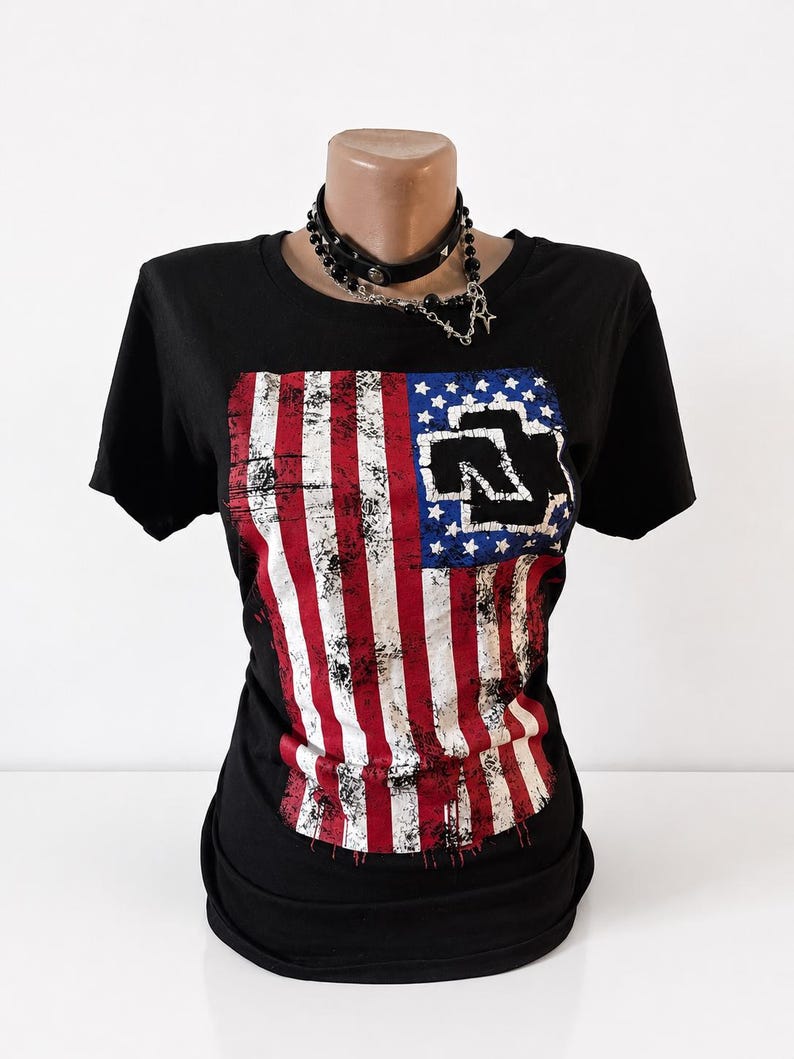 Puede incluir: Una camiseta negra de manga corta con un gr&aacute;fico de la bandera estadounidense desgastada en la parte delantera y el logotipo negro de Rammstein sobre el campo de estrellas azul. La prenda se muestra en un maniqu&iacute; con una gargantilla negra plateada.