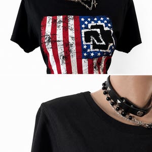 Puede incluir: Una camiseta negra de manga corta con un gr&aacute;fico de bandera estadounidense desgastada y el logotipo de Rammstein en negro superpuesto. La prenda se combina con una gargantilla de cuero negro decorada con tachuelas plateadas, cuentas y cadena.