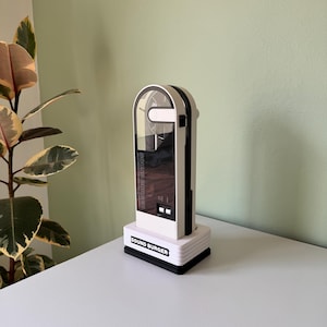 Peut inclure: Un appareil audio Sound Burger blanc et noir avec un couvercle en plastique transparent. L'appareil a une base rectangulaire et un sommet en forme d'arche. Les mots "SOUND BURGER" sont imprimés sur la base.