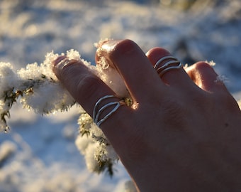 May include: A hand wearing silver rings with a wavy design. The rings are on a finger that is touching a snow-covered branch. The background is a blurred winter scene with sunlight.