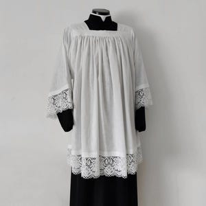 May include: A white liturgical surplice featuring a square neckline and gathered fabric that creates vertical pleats. The garment includes wide sleeves and a bottom hem decorated with intricate white lace trim, worn over a black cassock.