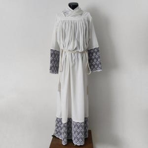 May include: A white liturgical garment with long sleeves and a floor-length skirt. The sleeves and hem feature a dark blue and white floral pattern. A rope belt cinches the waist, and the garment is displayed on a mannequin.