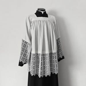 May include: A white surplice featuring a square neckline and long sleeves with intricate white floral lace trim at the cuffs and hem. The garment is gathered at the yoke and is displayed over a black cassock with a high collar and button closure.