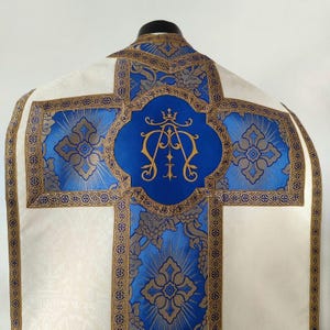 Może przedstawiać: Biała szata liturgiczna z wzorem krzyża w kolorze niebieskim i złotym. W centrum krzyża znajduje się niebieski owal z złotym monogramem "M.A." i koroną.
