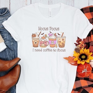 Peut inclure: T-shirt blanc avec un graphique de quatre tasses de café avec des décorations d'Halloween et le texte "Hocus Pocus I need coffee to focus".