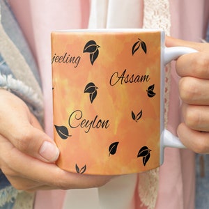 Può includere: Una tazza in ceramica bianca con manico. La tazza ha uno sfondo acquerellato arancione e giallo con disegni di foglie nere e le parole "feeling", "Assam" e "Ceylon" scritte in nero.