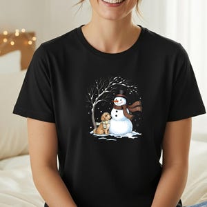 May include: Black t-shirt featuring a winter scene with a snowman, a dog, and a tree. The snowman wears a black top hat and a brown scarf. The dog is wearing a scarf. The background shows a snowy landscape.