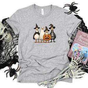 May include: A light gray t-shirt featuring a Halloween-themed graphic. The graphic depicts four cartoon geese dressed as a witch, a ghost, a pumpkin, and a skeleton. The shirt is surrounded by Halloween decorations, including a skull, spiders, and a book.