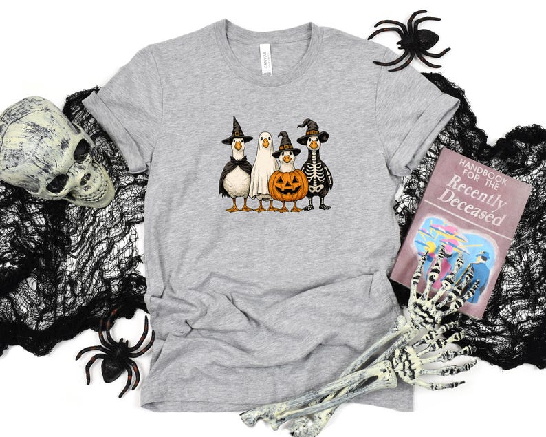 May include: A light gray t-shirt featuring a Halloween-themed graphic. The graphic depicts four cartoon geese dressed as a witch, a ghost, a pumpkin, and a skeleton. The shirt is surrounded by Halloween decorations, including a skull, spiders, and a book.