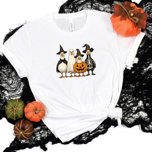 May include: White t-shirt featuring a Halloween-themed graphic. The design includes four cartoon ducks dressed in costumes: a witch, a ghost, a pumpkin, and a skeleton. The shirt is styled with a knot at the bottom and is set against a black, textured backdrop.