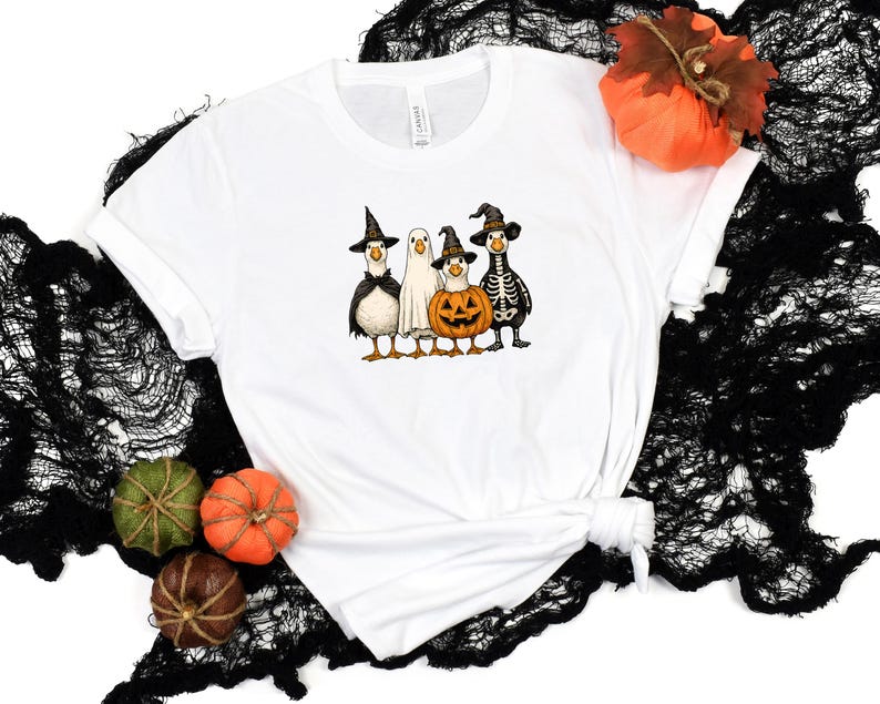 May include: White t-shirt featuring a Halloween-themed graphic. The design includes four cartoon ducks dressed in costumes: a witch, a ghost, a pumpkin, and a skeleton. The shirt is styled with a knot at the bottom and is set against a black, textured backdrop.