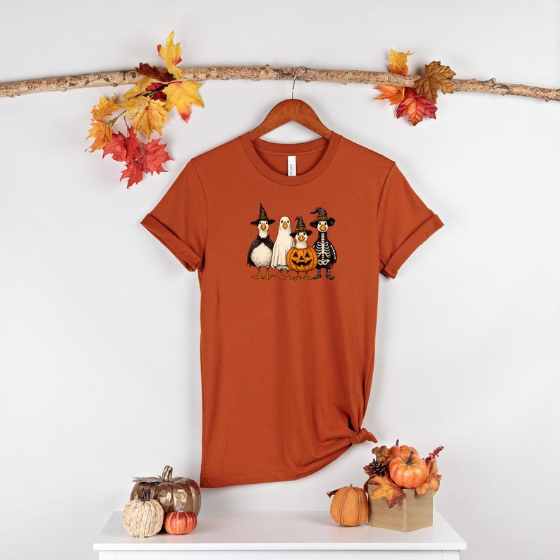 May include: Rust-colored t-shirt featuring a Halloween-themed graphic. The graphic depicts a penguin, a ghost, a jack-o'-lantern, and a skeleton, all wearing witch hats. The shirt is hanging on a wooden hanger.