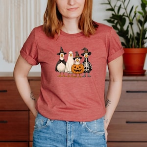 May include: A red t-shirt featuring a Halloween-themed graphic. The graphic depicts four cartoon ducks dressed in costumes: a witch, a ghost, a jack-o'-lantern, and a skeleton. The ducks are standing in a row.