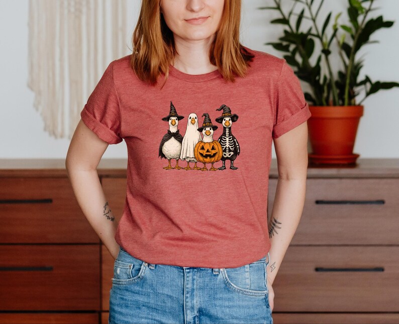 May include: A red t-shirt featuring a Halloween-themed graphic. The graphic depicts four cartoon ducks dressed in costumes: a witch, a ghost, a jack-o'-lantern, and a skeleton. The ducks are standing in a row.