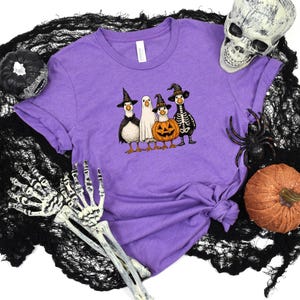 May include: A purple t-shirt featuring a Halloween-themed graphic of four cartoon ducks dressed in costumes: a witch, a ghost, a jack-o'-lantern, and a skeleton. The shirt is surrounded by Halloween decorations, including a skull, spider, and pumpkins.