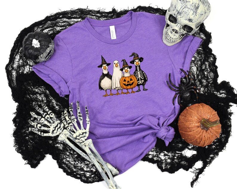 May include: A purple t-shirt featuring a Halloween-themed graphic of four cartoon ducks dressed in costumes: a witch, a ghost, a jack-o'-lantern, and a skeleton. The shirt is surrounded by Halloween decorations, including a skull, spider, and pumpkins.
