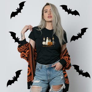May include: Black t-shirt featuring a Halloween-themed graphic of a pumpkin, ghost, and other characters. The model is wearing a black and orange patterned cardigan and ripped blue jeans. Black bat silhouettes are in the background.