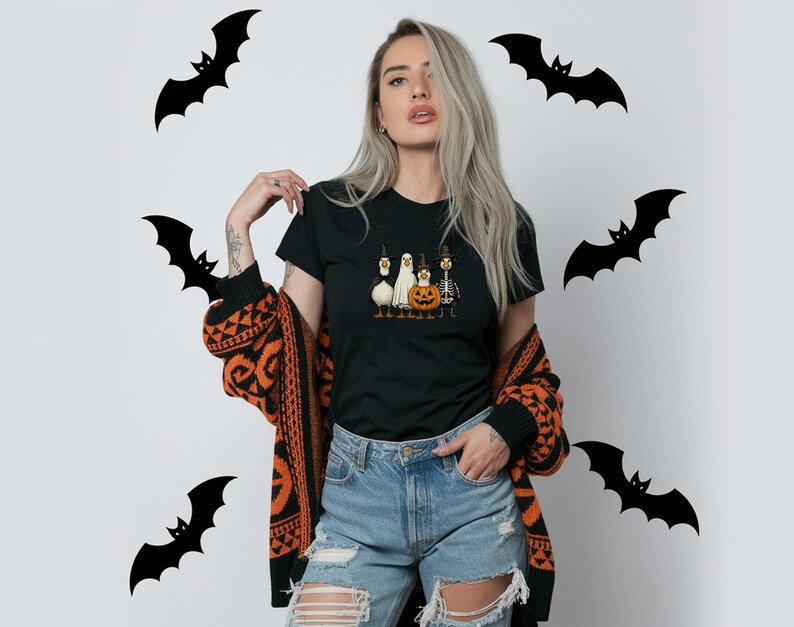 May include: Black t-shirt featuring a Halloween-themed graphic of a pumpkin, ghost, and other characters. The model is wearing a black and orange patterned cardigan and ripped blue jeans. Black bat silhouettes are in the background.