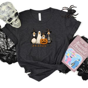 May include: Dark gray t-shirt featuring a Halloween-themed design with four cartoon ducks dressed as a witch, ghost, jack-o'-lantern, and skeleton. A decorative skull, spider, and a book titled "Handbook for the Recently Deceased" are also present.
