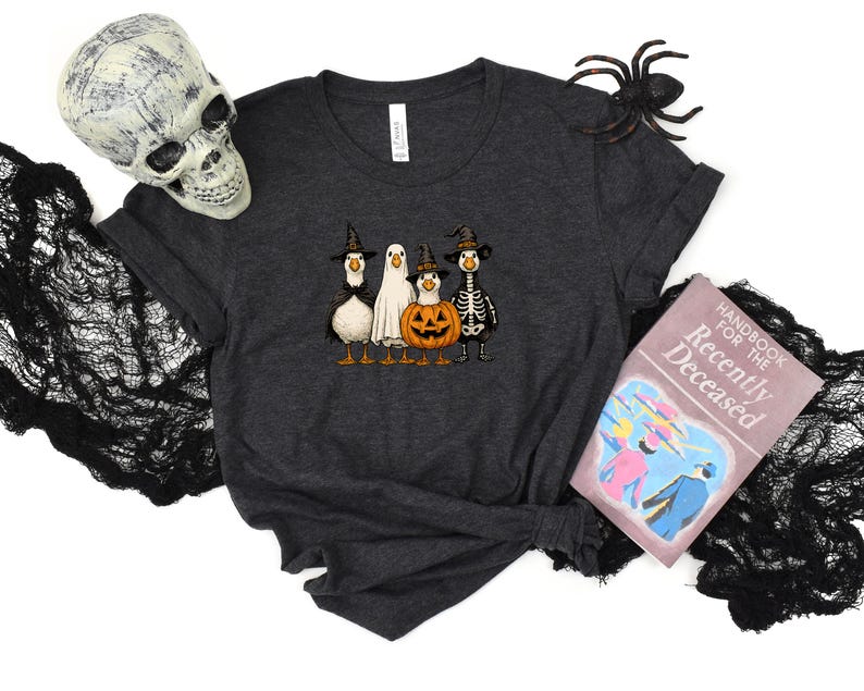 May include: Dark gray t-shirt featuring a Halloween-themed design with four cartoon ducks dressed as a witch, ghost, jack-o'-lantern, and skeleton. A decorative skull, spider, and a book titled "Handbook for the Recently Deceased" are also present.
