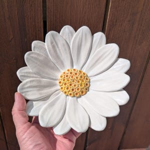 Puede incluir: Una gran margarita de cerámica blanca con un centro amarillo y naranja. La flor se sostiene en una mano, con un fondo de madera. Los pétalos son blancos y el centro tiene un diseño texturizado.