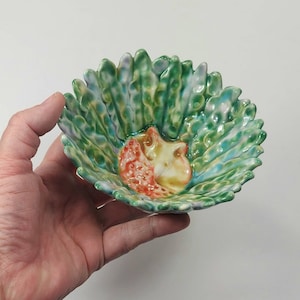 Puede incluir: Un pequeño cuenco de cerámica con un diseño de hojas verdes, azules y blancas. El cuenco tiene un diseño de zorro naranja y amarillo en relieve en el centro.