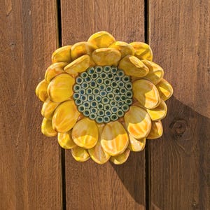 May include: A ceramic sunflower wall hanging with yellow petals and a blue-green center. The sunflower is mounted on a wooden surface, creating a rustic and decorative piece for home decor.