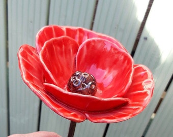 May include: A red ceramic poppy flower with a brown centre. The flower is on a black stem.