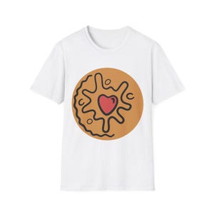 Puede incluir: Camiseta blanca con un diseño de galleta. La galleta es marrón con un contorno negro y un corazón rojo en el centro. El diseño es de estilo caricatura y está impreso en la parte delantera de la camiseta.