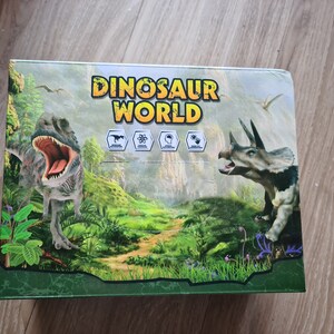 Könnte beinhalten: Eine grüne und braune Box mit den Worten "Dinosaur World" auf der Vorderseite. Die Box zeigt einen grünen, braunen und grauen Dinosaurier mit offenem Maul und einen grünen, braunen und grauen Triceratops mit einem Horn auf der Nase. Die Box hat einen grünen Hintergrund mit einer Dschungelszene.