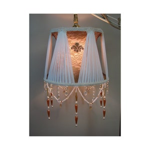 May include: A white lampshade with a lace design and beaded fringe. The lampshade is illuminated from within, casting a warm glow.