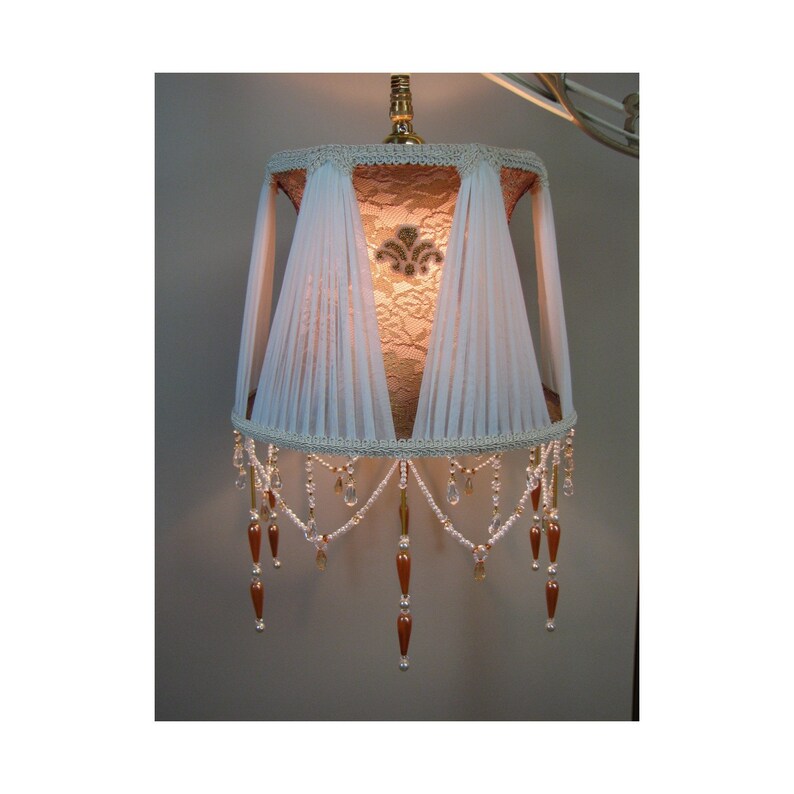 May include: A white lampshade with a lace design and beaded fringe. The lampshade is illuminated from within, casting a warm glow.