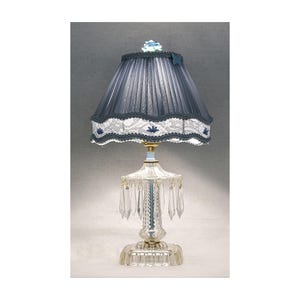May include: A vintage-style table lamp with a pleated navy blue lampshade, trimmed with white lace and blue floral accents. The lamp features a clear glass base with hanging crystal accents and a light blue and gold stem. A small blue flower decorates the top of the lampshade.