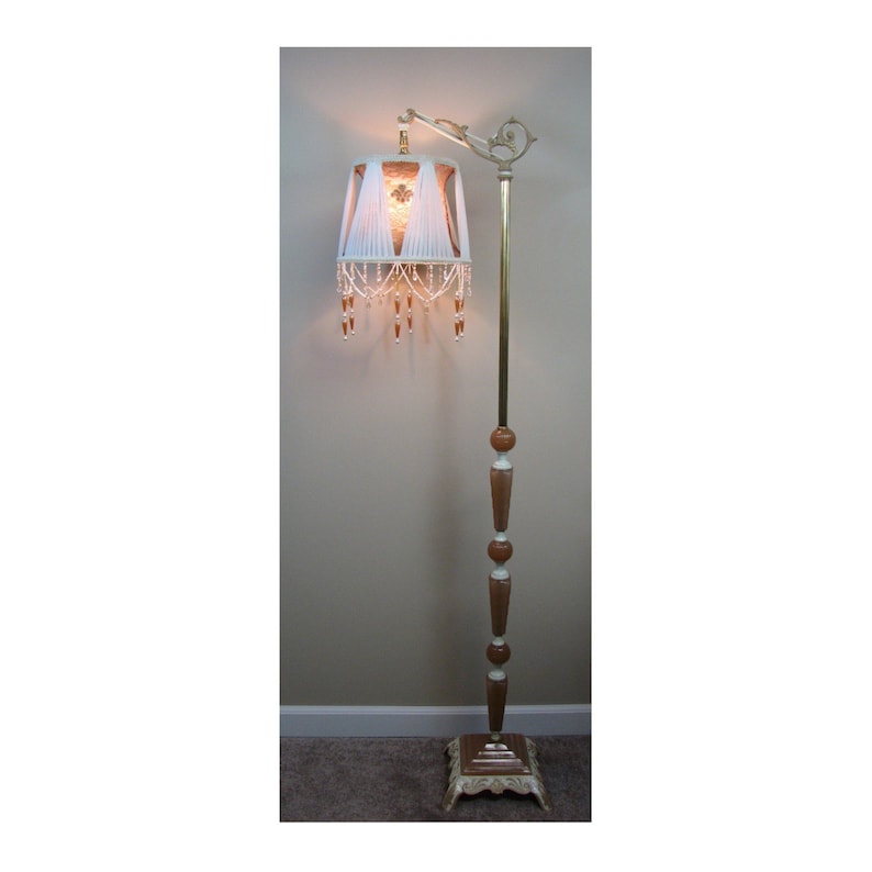 May include: A white floor lamp with a beaded fringe shade and a brown wooden base. The lamp has a gold metal arm and a white and gold base.
