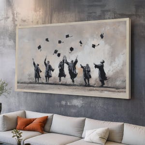 以下が含まれることがあります： 卒業シーンを描いた額入りのプリント。卒業ガウンを着たシルエットが、帽子を空中に投げながらジャンプしています。アートワークはグレースケールで、ニュートラルなトーンの背景です。