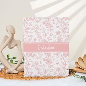 May include: A notebook featuring a pink floral design and the name "Sebastian" on a pink band. The notebook is displayed on a white surface, with a decorative sculpture and an open book visible in the background.