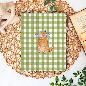 May include: A notebook with a green and white gingham pattern. The cover features a cartoon orange tabby cat holding two fish. The notebook is on a woven placemat with a decorative sculpture and a book in the background.