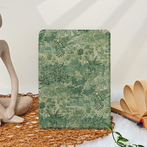 May include: A rectangular tablet case with a green and white toile design showing a landscape with trees and foliage. The case is on a woven mat, with a sculpture and open book in the background.