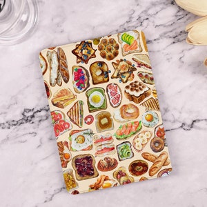 May include: A rectangular tablet case featuring a repeating watercolor illustration of various breakfast foods including toast with toppings, fried eggs, bagels, croissants, sliced fruit, bacon, and assorted breads on a light cream background.
