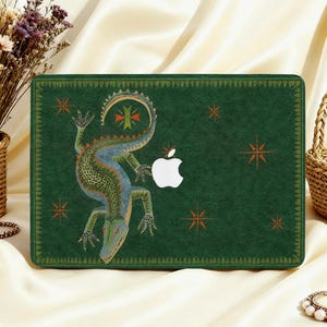 Può includere: Un adesivo per laptop verde scuro con un'illustrazione dettagliata di una lucertola con squame strutturate, circondata da piccoli simboli a forma di stella arancione e un bordo decorativo. Il logo Apple bianco è visibile al centro della cover.