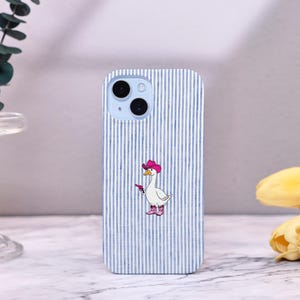 Może przedstawiać: Etui na telefon z niebiesko-białym wzorem w paski. Etui przedstawia kresk&oacute;wkową gęś w r&oacute;żowym kapeluszu kowbojskim i butach, trzymającą pistolet. Etui na telefon ma jasno niebieski moduł aparatu.